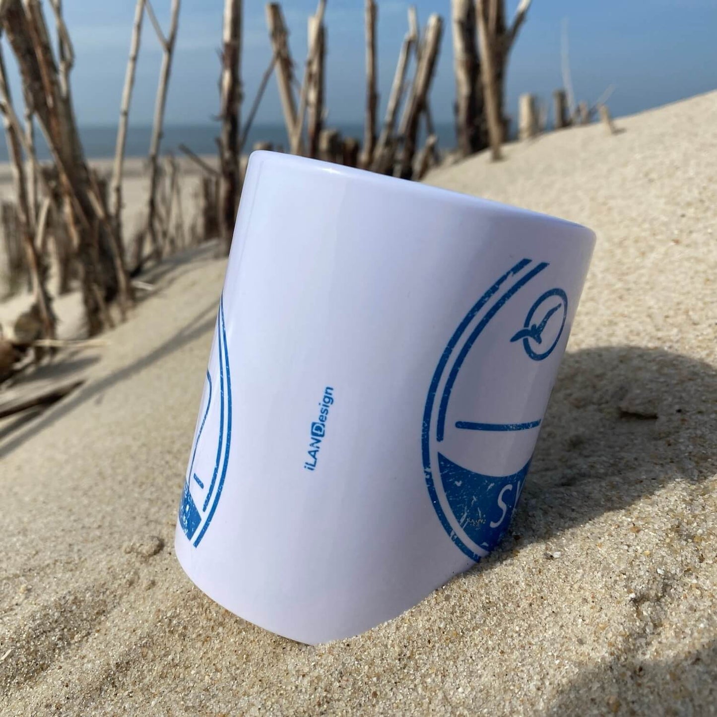 Weißer Becher liegt am Strand auf Sylt. Im Hintergrund ist das blaue Meer zu sehen. Der Becher ist in blau bedruckt mit dem Begriff Sylt, einem Strandkorb sowie einer Möwe die durch die Sonne fliegt.