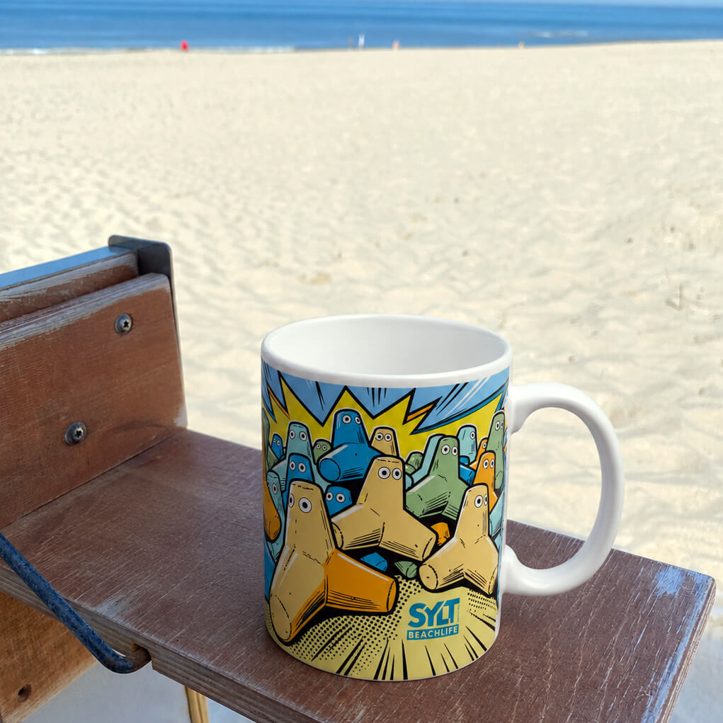 Sylt-Keramik-Becher mit Rundherum-Druck der Sylter Tetrapoden im Popart-Stil steht auf einem Strandkorbtischchen mit Blick auf den Strand und die Nordsee.