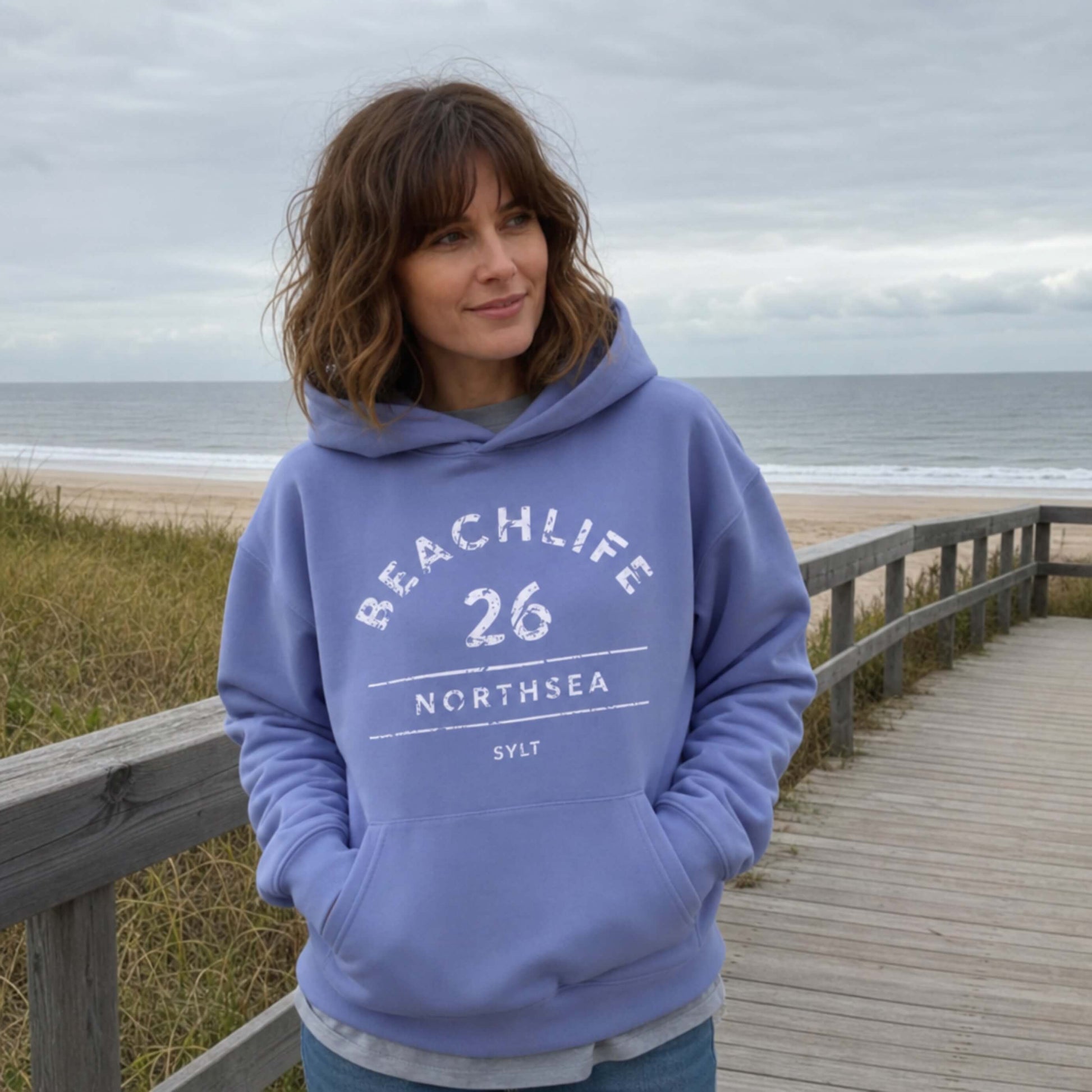 Braunhaarige Frau in violetem Hoodie mit "BEACHLIFE 26 NORTHSEA SYLT" Aufdruck in entspannter Strandszenerie.