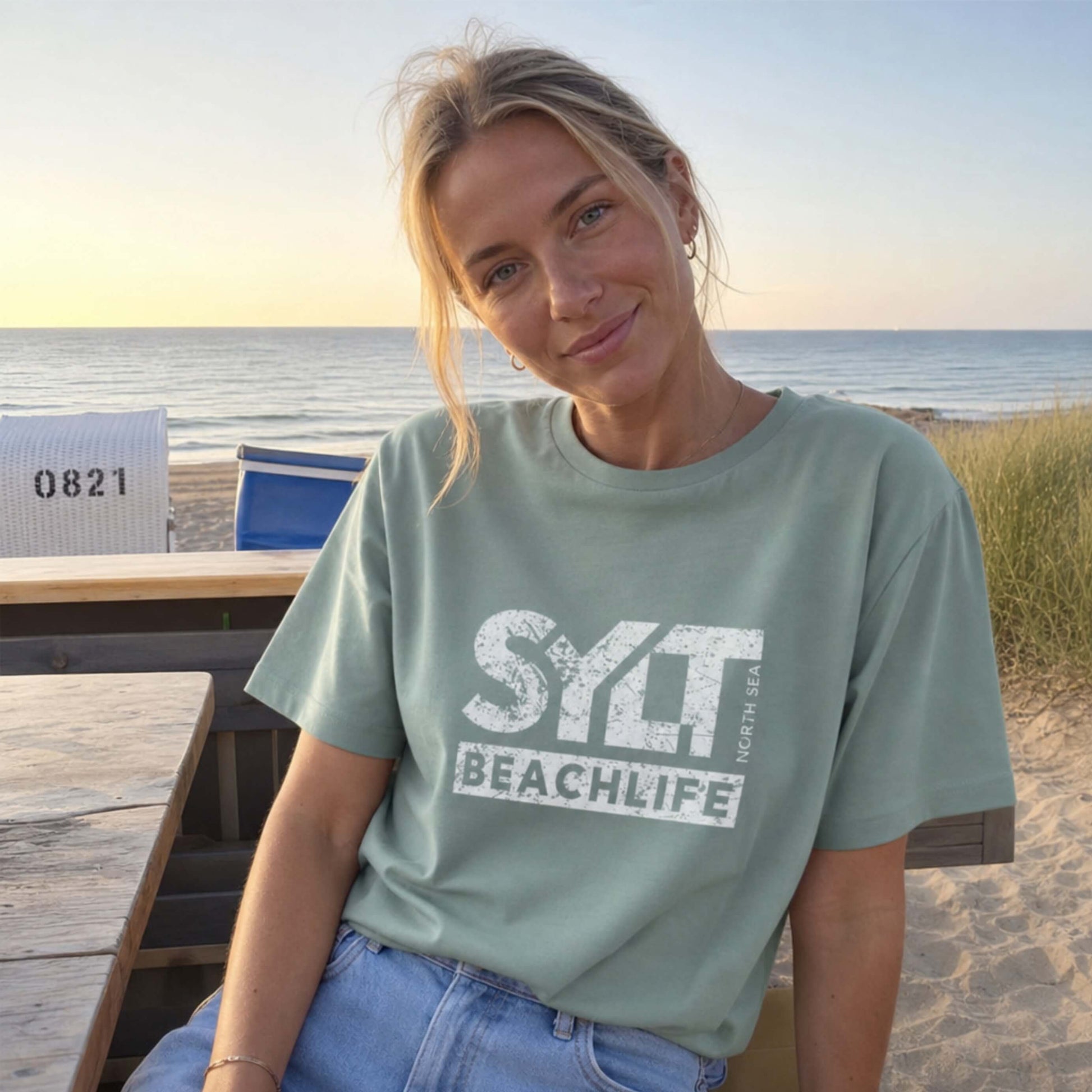 Frau in aloe farbenden Sylt T-Shirt steht auf sylt. Im Hintergrund sieht man Dünen, Strandkörbe am Strand und die Nordsee.