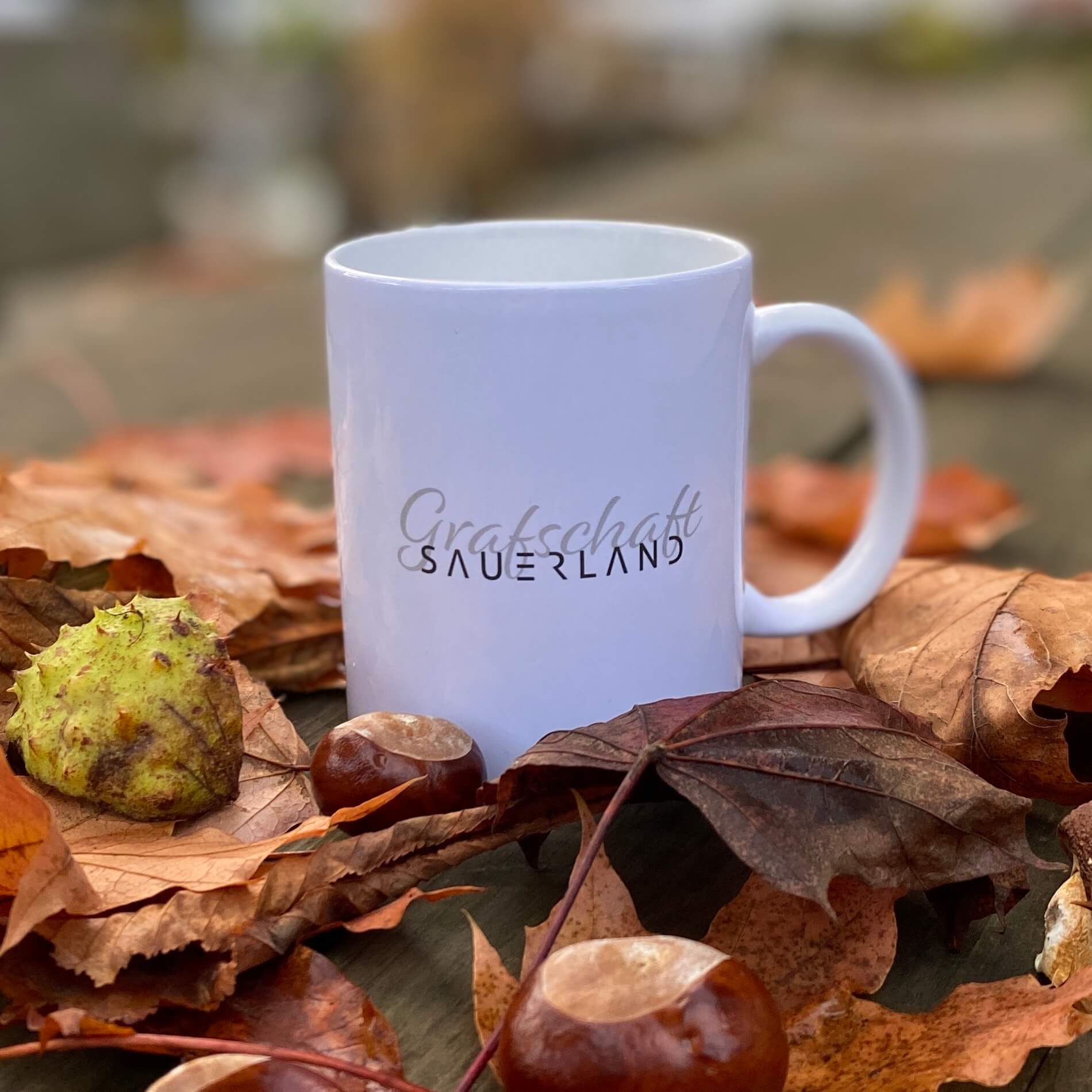 Weiße Kaffee- oder Tee-Tasse mit dem Dorfnamen Grafschaft in geschwungener Schrift sowie mit dem New Sauerland Schriftzug beschriftet. Der Becher steht auf einem Holztisch mit herbstlichem Laub und Kastanien.