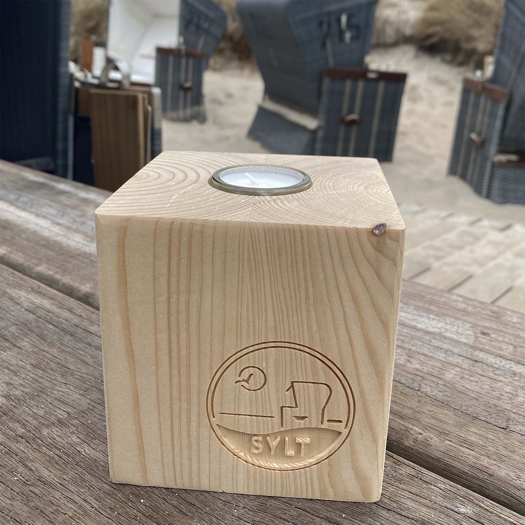 Holz Teelichtwürfel steht in Strandbistro auf einem rustikalen Holztisch. Im Hintergrund sind Strandkörbe zu sehen. Der Teelichtwürfel hat eine Fräsung mit dem Begriff Sylt, einem Strandkorb sowie einer Möwe, die durch die Sonne fliegt. 